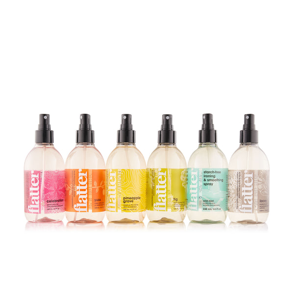 Six clear plastic spray bottles of flatter hair product lined up on a white background, featuring pink, orange, yellow, green, and gray labels.