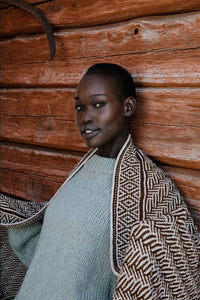 picture of a person leaned against a wooden wall and has a knit shawl wrapped around them