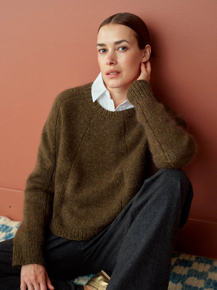 Woman wearing a brown sweater and white shirt against a pink wall.