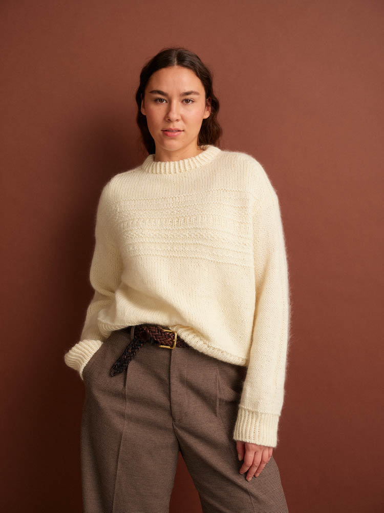 Woman wearing a cream sweater and brown pants against a brown background