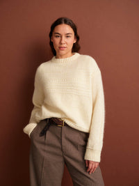 Woman wearing a cream sweater and brown pants against a brown background