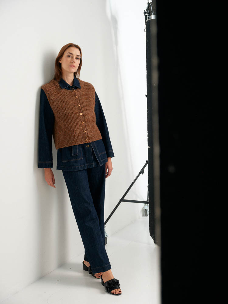 Woman wearing a brown vest over a black top and jeans, standing against a white background.