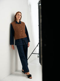 Woman wearing a brown vest over a black top and jeans, standing against a white background.