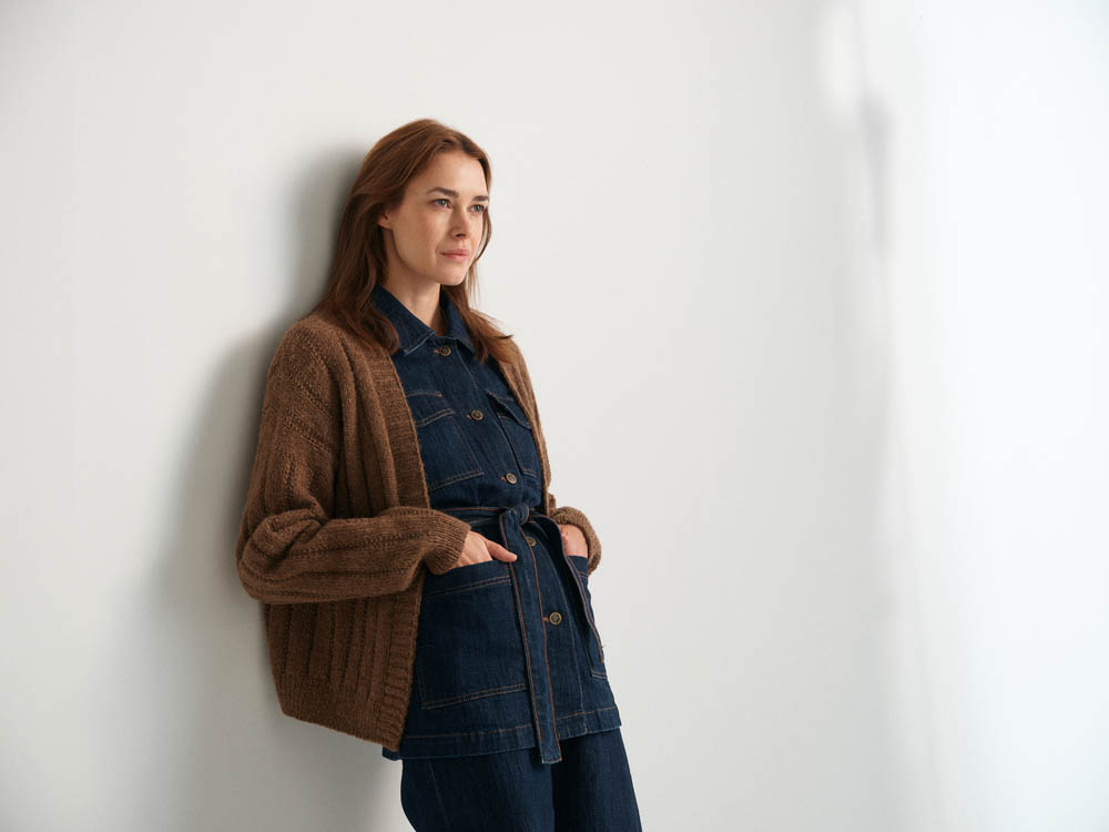 Woman wearing a brown cardigan over a denim outfit against a white wall