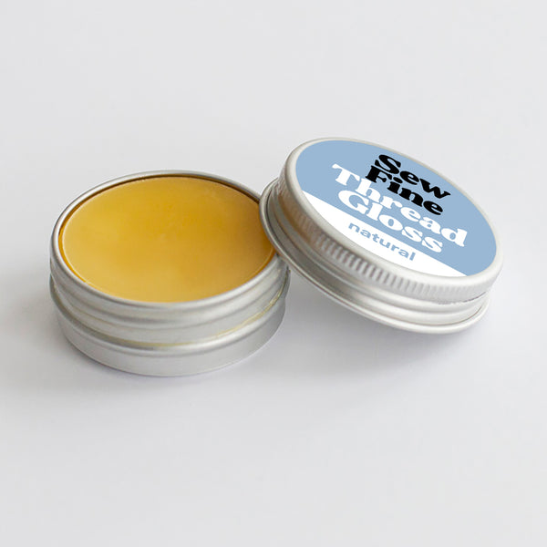 An open round silver tin of Sew Fine Thread Gloss showing the yellow wax balm inside, with the lid leaning against it. The lid has a light blue label with white and black text reading 'Sew Fine Thread Gloss natural'.