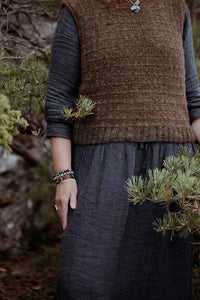 Person wearing a brown patterned vest and dark skirt standing among greenery.