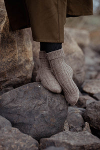 A close-up of a person standing on large rocks outdoors, wearing ribbed knit ankle socks in a taupe-beige color and dark pants.