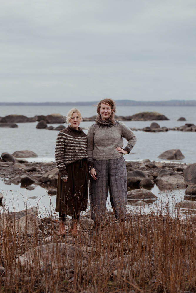 Two women standing on a rocky beach with a body of water and sky in the background.