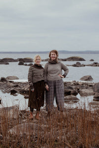 Two women standing on a rocky beach with a body of water and sky in the background.