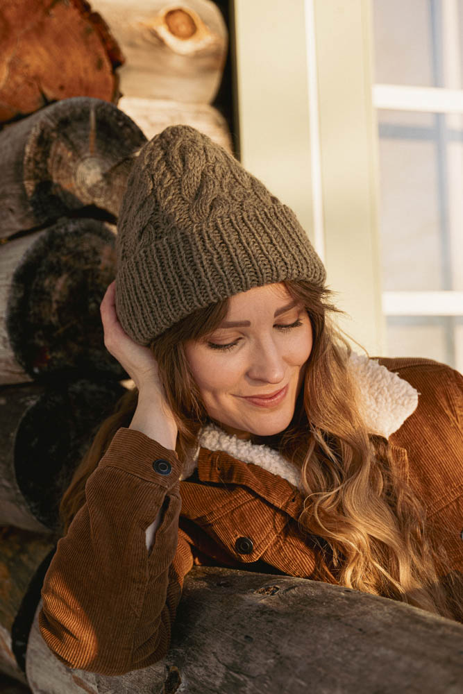 image of a knit hat being worn on a woman sitting outside by a log house.  