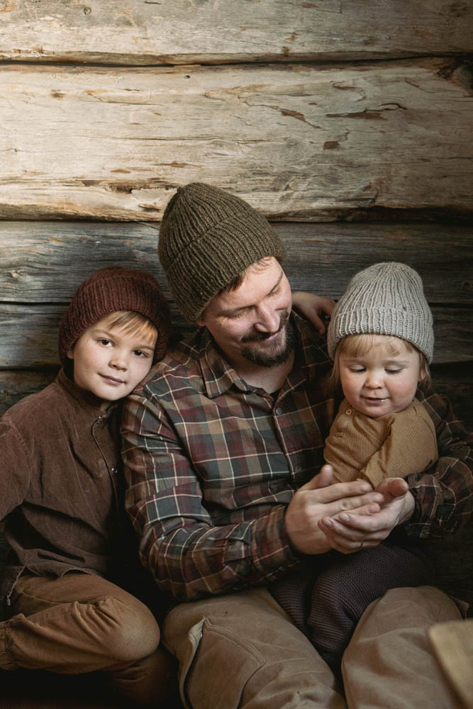 image of 2 children and 1 adult male wearing knit hats outside in front a log wall.