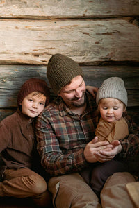 image of 2 children and 1 adult male wearing knit hats outside in front a log wall.