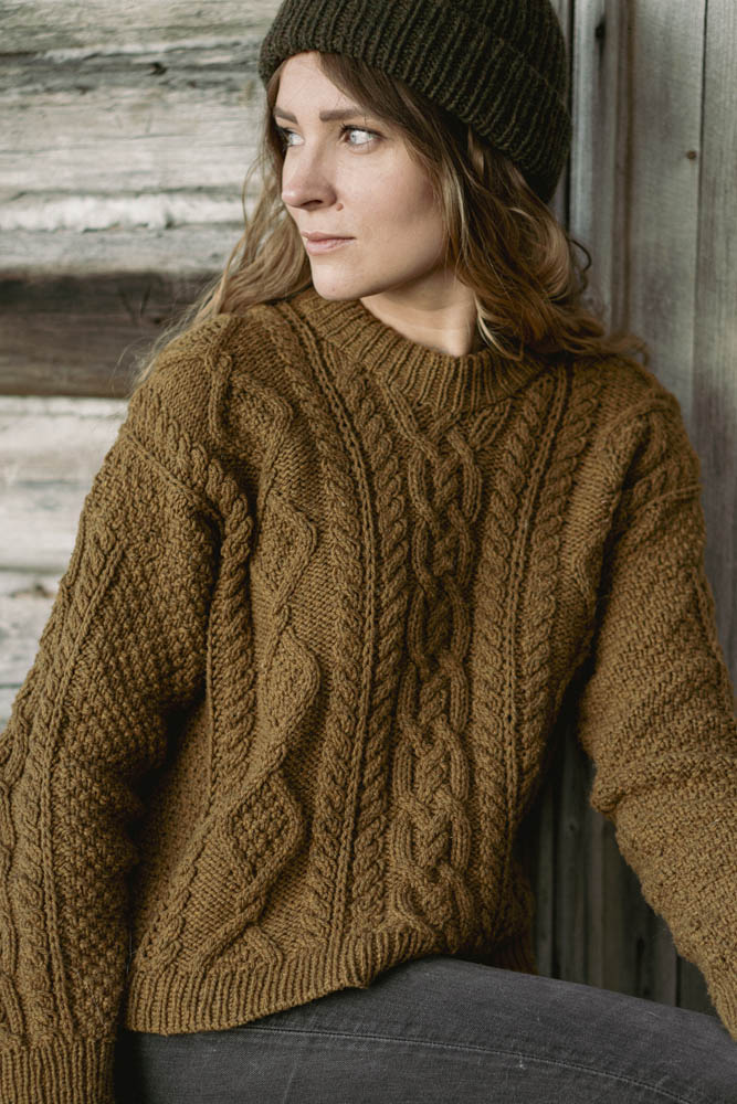 image of a woman wearing a cable knit sweater with a knit hat outside in front of wood wall